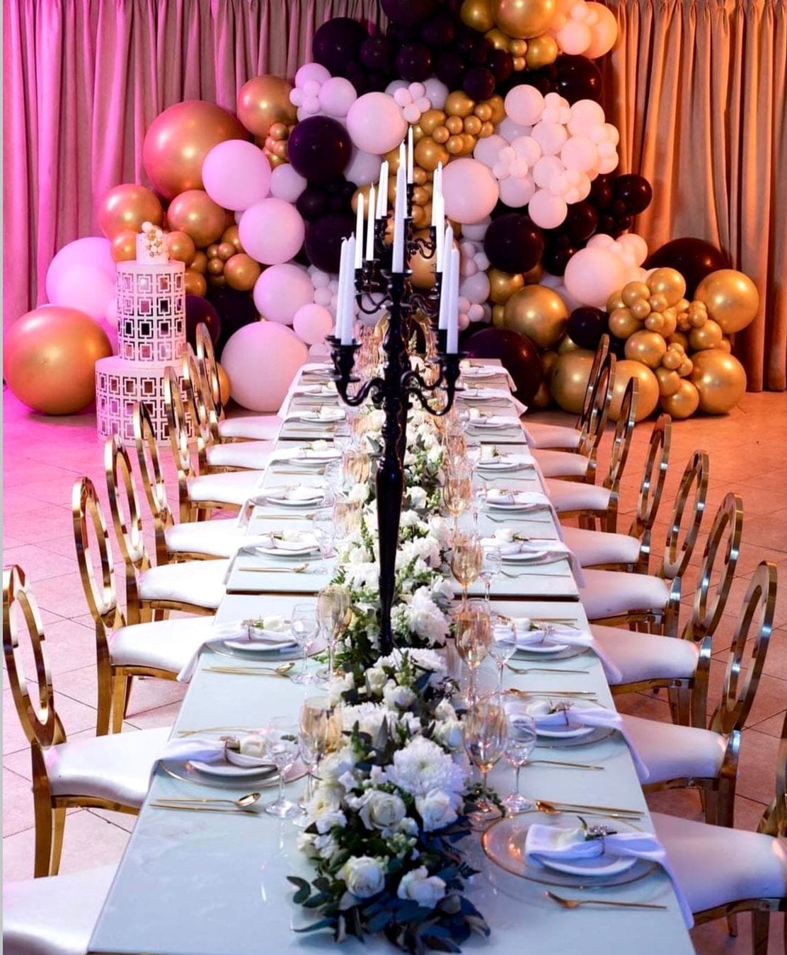 Dramatic long tablescape with burgundy, pink, and gold balloon arch, black candelabra, and gold oval-back chairs