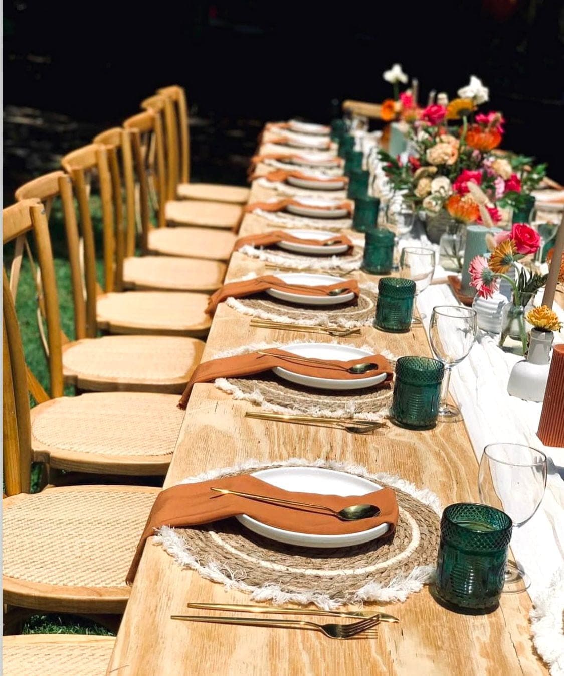 Rustic outdoor dinner on a raw wood table with jute placemats, clay napkins, bright florals, and wooden cross-back chairs