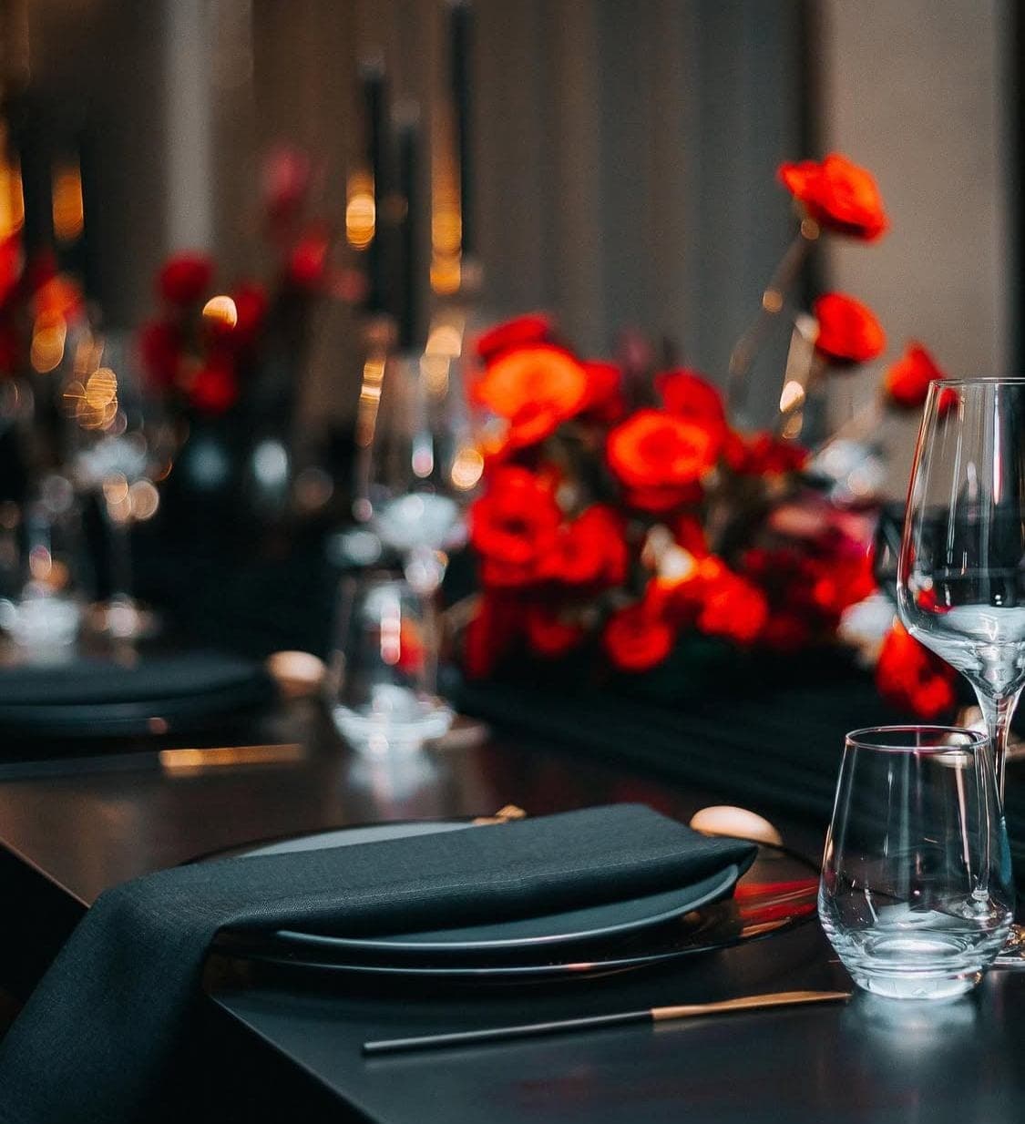 Moody black tablescape close-up with vivid red ranunculus centerpieces, black napkins, and candlelight