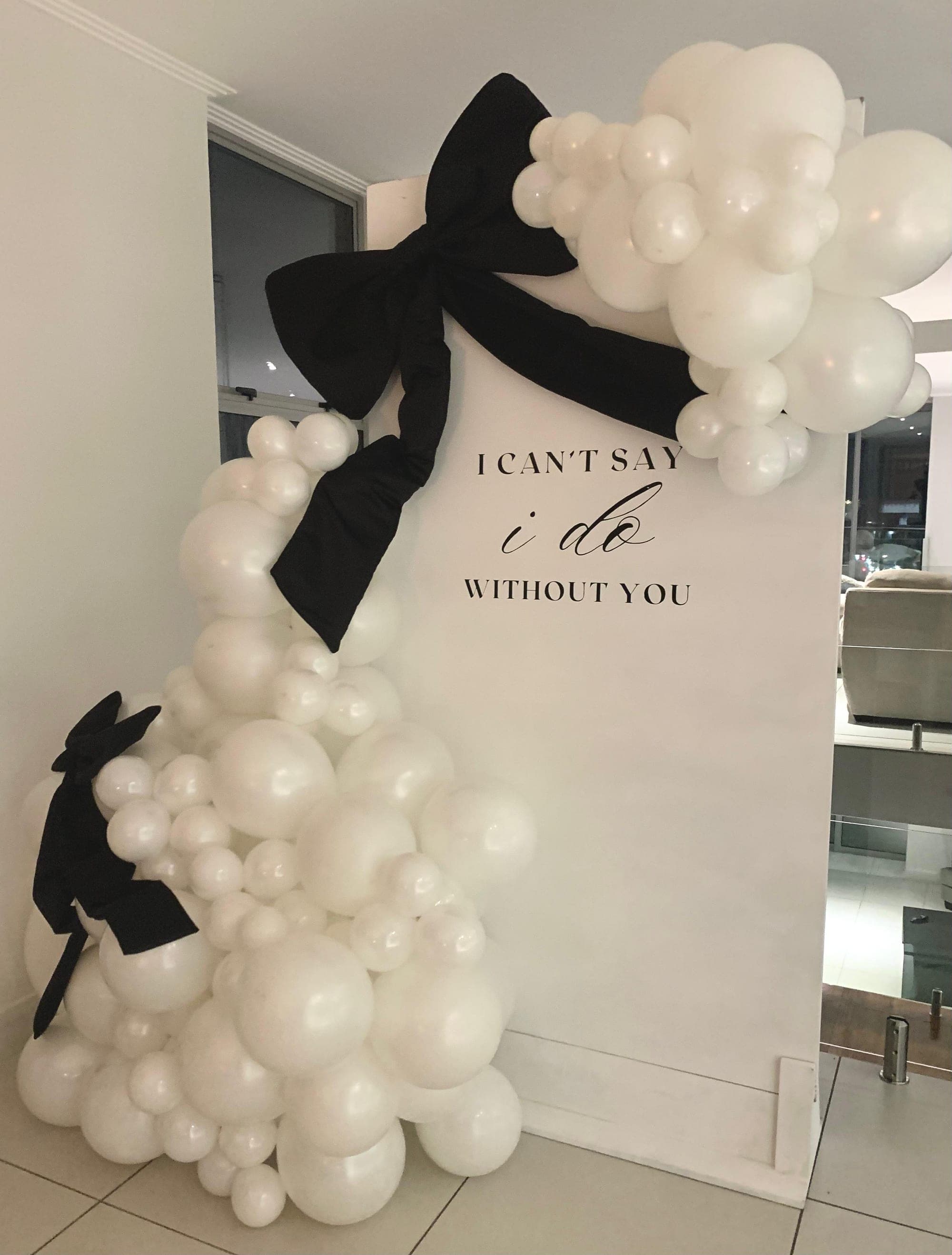 White balloon arch with black bows and 'I can't say I do without you' bridesmaid proposal sign by Frontrow Creatives